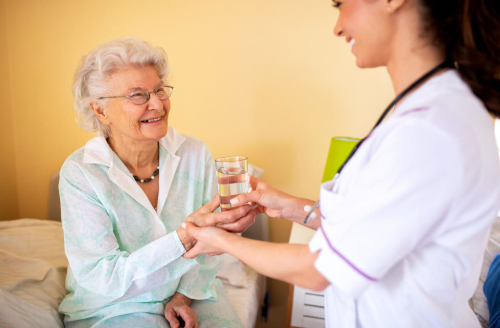 A lady nurse brings water to an elderly woman in her bed at a assisted living facility.