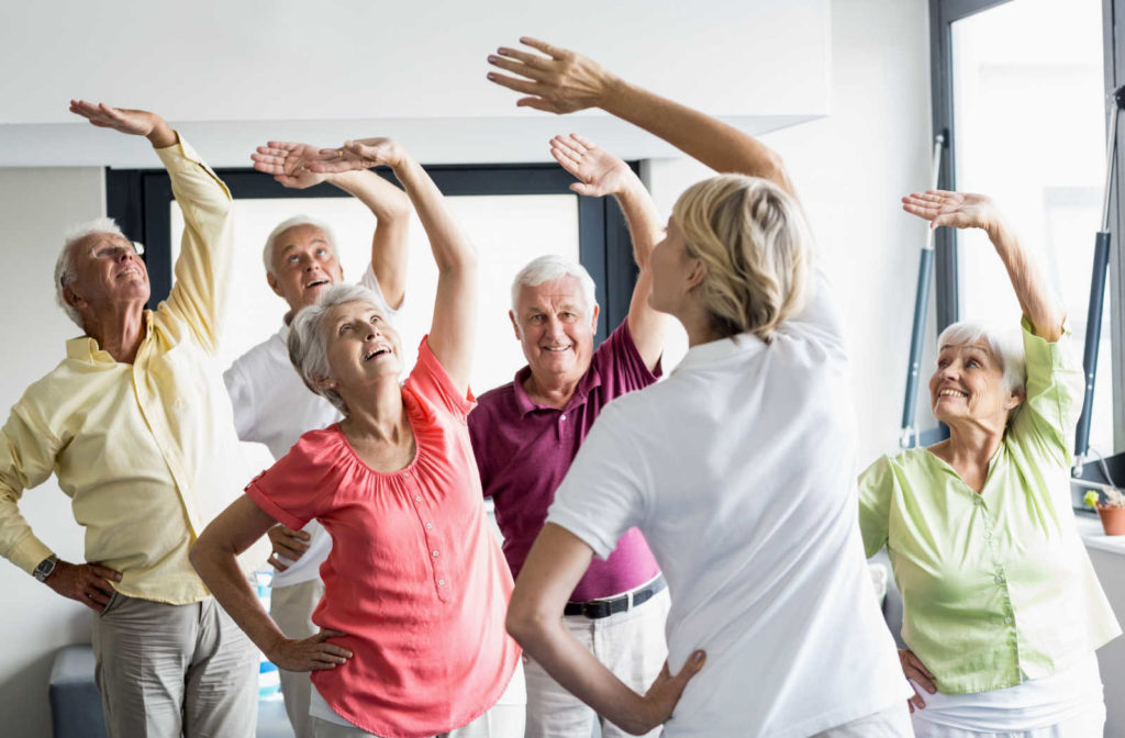 A group of seniors in a senior living facility sitting and exercising together under the supervision of a trained staff.
