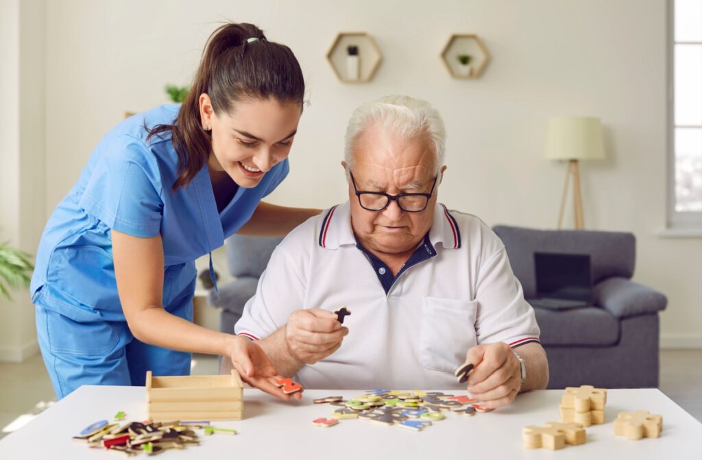 An older adult works on a puzzle at a table while a caregiver in a blue uniform provides friendly support in a bright and welcoming space.