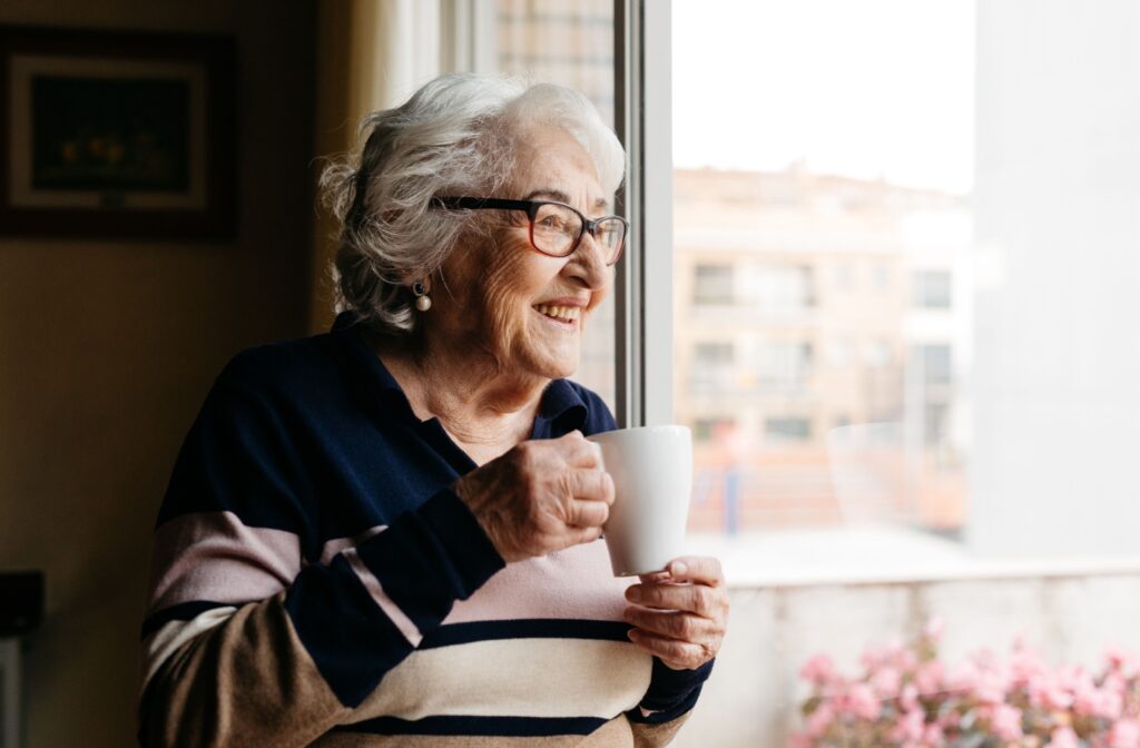 A senior holding a white mug smiles thoughtfully as they look out the window in their assisted living community.