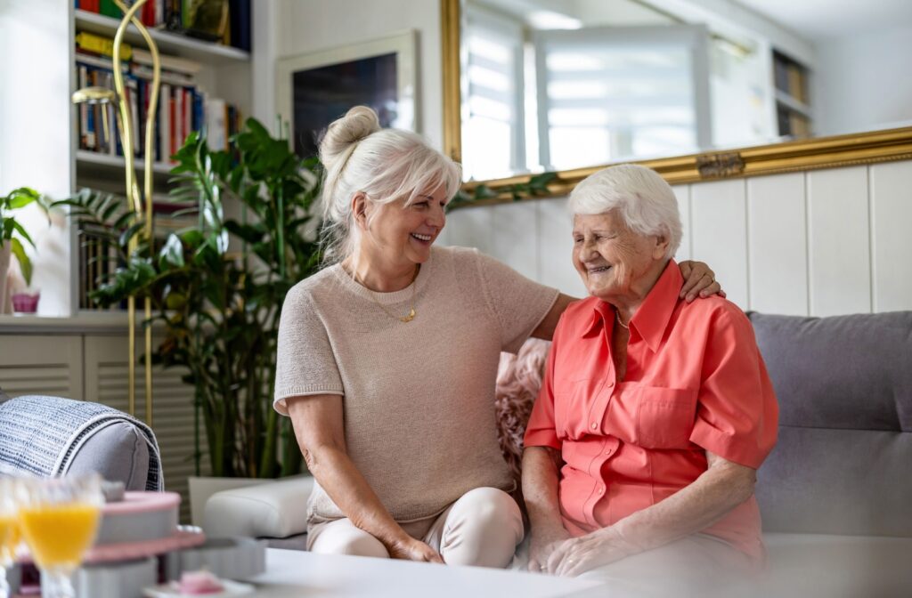 An older adult sits on a grey couch in their private apartment. Their adult child wraps an arm around their shoulders.