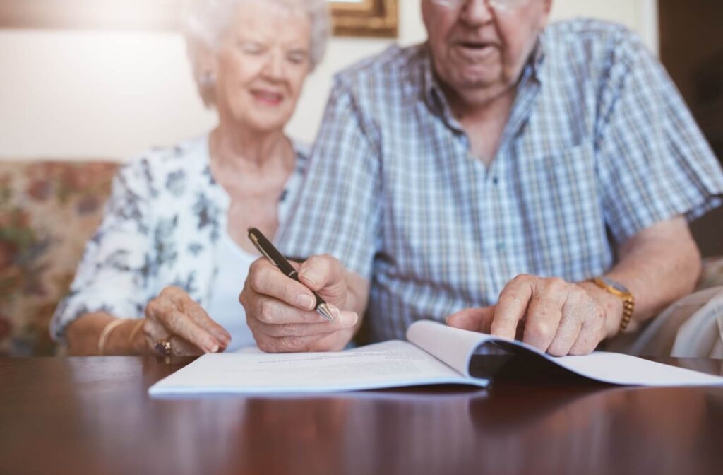 An older adult signs an application form for senior living while their smiling spouse watches